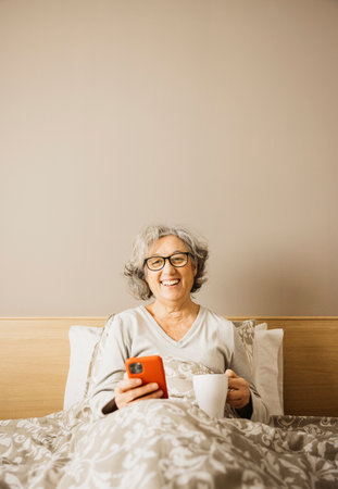 Portrait of a senior woman sitting on bed in the morning with pajama and drinking coffee with a white mug while checking her phone.の写真素材