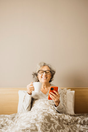 Senior woman sitting on bed in the morning with pajama and drinking coffee with a white mug while checking her phone.の写真素材