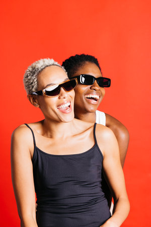 Portrait of a cheerful young female couple, wearing sunglasses, together in front of a red background at studio. Two women together in studio.の写真素材