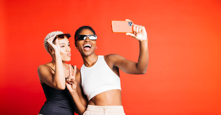 Portrait of a cheerful young female couple, wearing sunglasses and taking a selfie in front of a red background at studio. Two playful women together in studio.の写真素材