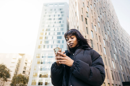 Young beautiful black woman wearing black clothes typing an online message on her phone while walking on the street of a city.の写真素材