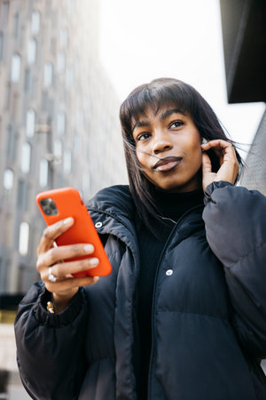Woman walking in the city with wireless headphones and a phone. Young beautiful woman wearing black clothes using headphones and a phone while walking on a city.の写真素材