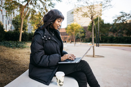 Young beautiful woman using sitting on a public park with a laptop. Young black woman working on the street with a laptop.の写真素材