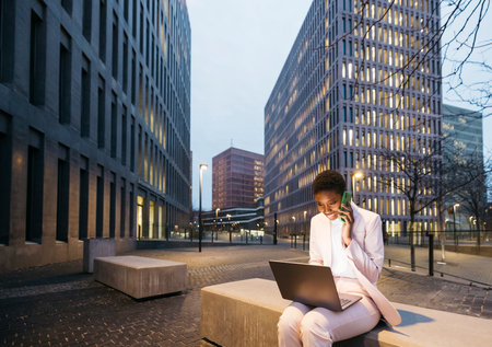 Young stylish businesswoman sitting on a street bench talking on a green phone while working on a laptop in street in city. Young woman working on the street with her laptop in the evening.の写真素材