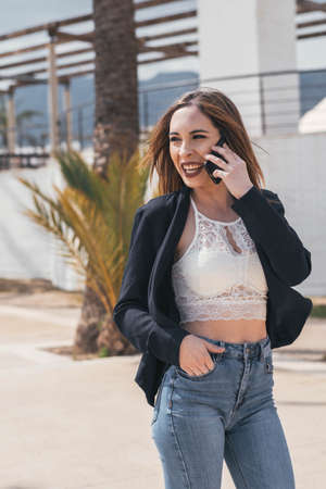business woman smiles while talking on the mobile phone and looks to the side. She wears a dark blazer and jeans.の写真素材