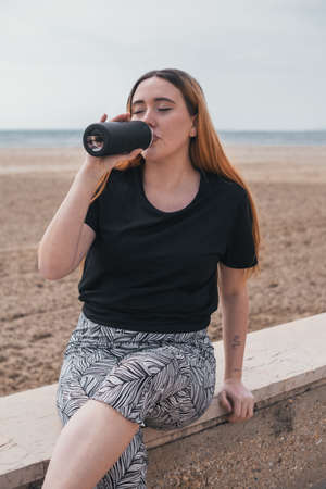 redhead runner girl sitting by the beach taking a break while drinking water from a black bottleの写真素材