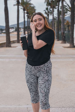 sporty redhead girl smiles with a black bottle in her hand in a beach environment with palm treesの写真素材