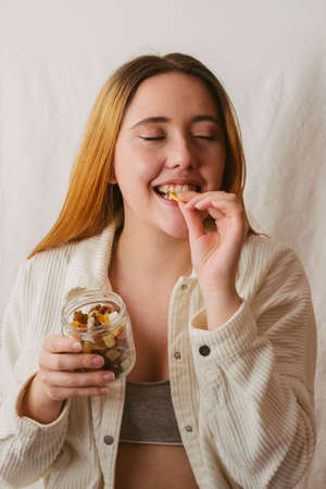 red-haired girl smiling while biting a piece of dried banana and holding a glass jar full of dried...
