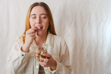 red-haired girl eating a piece of dried coconut while holding a glass jar full of dried fruitの写真素材