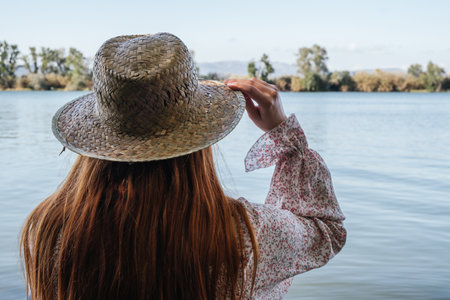woman from behind touching her straw hat with one hand in front of a riverの写真素材