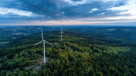An aerial perspective showcases wind turbines amid a forest setting under a cloudy sky. The composition emphasizes the turbines' height and integration with the landscape, with varying shades of green and blue. This image might be suitable for illustrating topics related to renewable resources or ecological concepts.の素材
