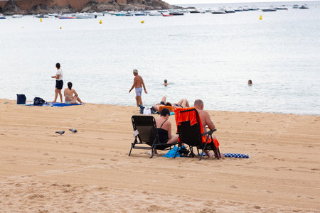 Tourists are enjoying a sunny day at the beach, relaxing under umbrellas with a sailboat visible in...