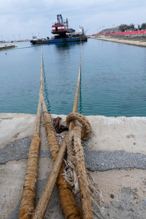 Mooring ropes and rusty chain securing a crane barge in a harbor, with construction site in backgroundの写真素材