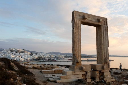 The ancient Portara, a marble doorway, stands on the islet of Palatia in Naxos, Greece, offering stunning views of the Aegean Sea and the town of Chora at sunsetの写真素材