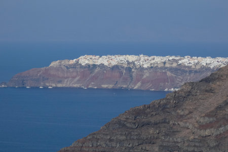 White imerovigli town on santorini island in daylight rising above colorful caldera cliffs with blue sea and dark volcanic rocks in foregroundの写真素材