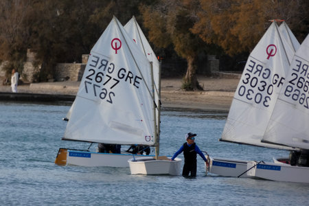 Young sailors are competing in a regatta, navigating their small sailing boats on the blue waters of the Aegean Sea, while coaches observe from a nearby yachtの写真素材