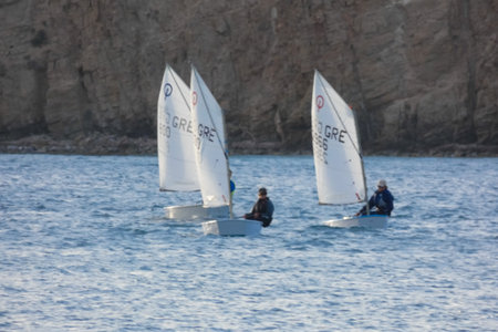 Young sailors are competing in a regatta, navigating their small sailing boats on the blue waters of the Aegean Sea, while coaches observe from a nearby yachtの写真素材