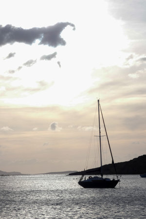 Peaceful scene of a sailboat at sunset, with calm water reflecting the sky and cloudsの写真素材