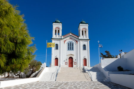 Traditional white church with red doors, byzantine flag, and greek flag waving in the wind, under a deep blue skyの写真素材