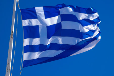 National flag of Greece waving on flagpole on sunny day with blue sky and some cloudsの写真素材