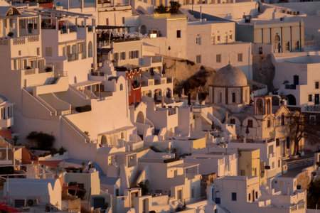 The white dome of an orthodox church rises above the rooftops of fira, santorini, bathed in the warm glow of the setting sunの写真素材