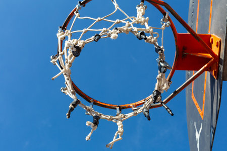 Close-up of an outdoor basketball hoop featuring a damaged net, set against a vibrant blue skyの写真素材