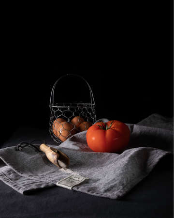 Vertical photograph of a beautiful composition of a fresh tomato and a basket with eggs accompanied by a spoon of pink salt and a gray cloth on a black background.の写真素材