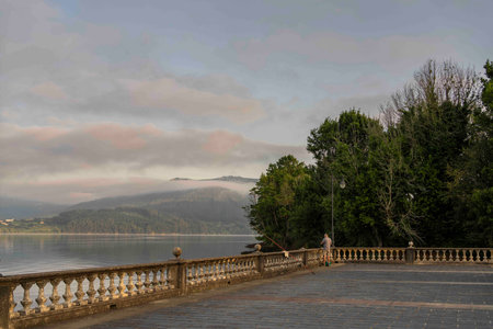A serene waterfront scene featuring a stone railing along a calm lake. A person is fishing near the edge, surrounded by lush greenery and misty mountains in the background. The sky is cloudy, creatingの写真素材