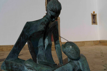 A bronze sculpture depicting a stylized figure of a parent holding a child, showing an emotional connection. The figures are abstract with elongated forms, set against a white wall, highlighting the artistry and craftsmanship.の写真素材
