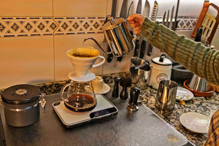 A person carefully pours hot water from a stainless steel kettle over coffee grounds using a pour-over coffee maker. The scene is set in a cozy kitchen, with various coffee utensils neatly arranged, highlighting the art of manual coffee brewing.の写真素材