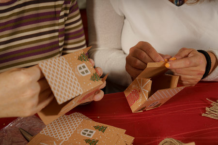 Two people assembling decorative gingerbread house boxes. One person holds a partially assembled box while the other is attaching a piece. The setting is festive with a red tablecloth and holiday decoの写真素材