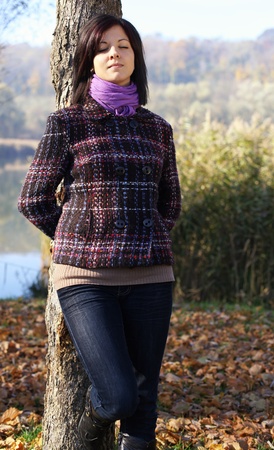 Outdoors portrait of happy young woman leaning to tree trunk in autumn forestの写真素材