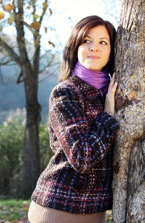 Outdoors portrait of happy young woman leaning to tree trunk in autumn forestの写真素材