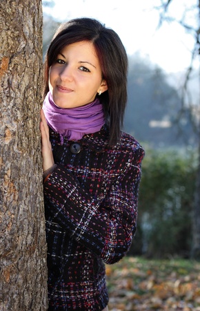 Outdoors portrait of happy young woman leaning to tree trunk in autumn forestの写真素材