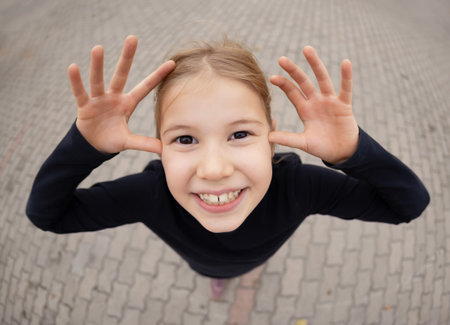 Playful young child making a funny face while smiling and laughing outdoors. Captured from a top view with a wide-angle perspective, this authentic lifestyle portrait shows joy, energy, and positive emotion. Natural daylight, casual clothing, and a street background create a candid, real-life moment of childhood happiness. Ideal for concepts related to joy, fun, emotions, parenting, lifestyle, and positive family themes.の写真素材