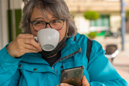 white haired woman drinking coffee at the bar while writing and looking at the phoneの写真素材