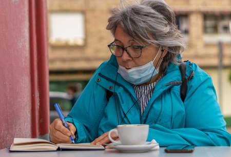 white haired woman drinking coffee at the bar while writing and looking at the phoneの写真素材