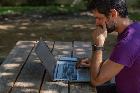 young man working at his computer on a wooden table in the park on a sunny dayの写真素材