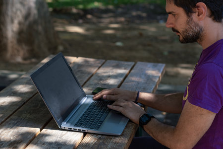 young man working at his computer on a wooden table in the park on a sunny day
