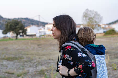 Young mother with her toddler kid boy on back in ergonomic baby carrier in autumn nature. Active mother conceptの写真素材