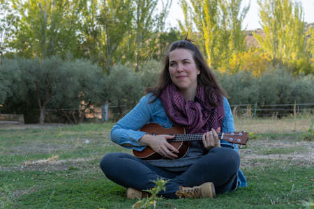 young brunette girl with long hair playing ukulele in the field on the grass