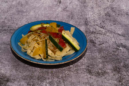 homemade vegan pasta with zucchini, onion, peppers and orange on a blue plate with a gray backgroundの写真素材