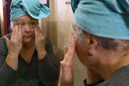 older woman creaming her face in her bathroom in a blue bathrobeの写真素材