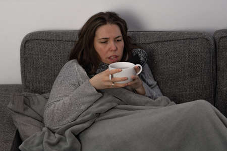 young brunette girl with long hair, sick lying on the sofa in her house taking medicine and an infusionの写真素材