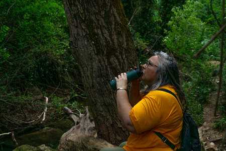 older white-haired woman drinking water from a bottle in a field sitting on a tree trunkの写真素材