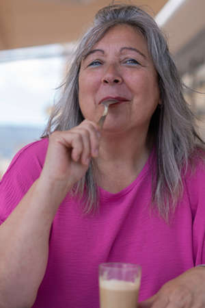 older white-haired woman in pink t-shirt drinking coffee in a bar sucking the spoon , funnyの写真素材