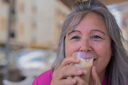 close-up of an older white-haired woman drinking coffee with milk in a bar looking at the camera...