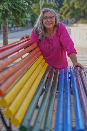 older woman with long white hair and glasses smiling leaning on a bench painted with the colors of the LGBT flag.の写真素材