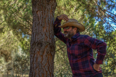 close-up of a man with a beard, hat and sunglasses leaning against a pine tree in the forestの写真素材