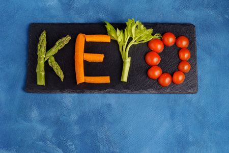 keto word drawn with asparagus, carrots, celery and cherries on a black board and blue backgroundの写真素材